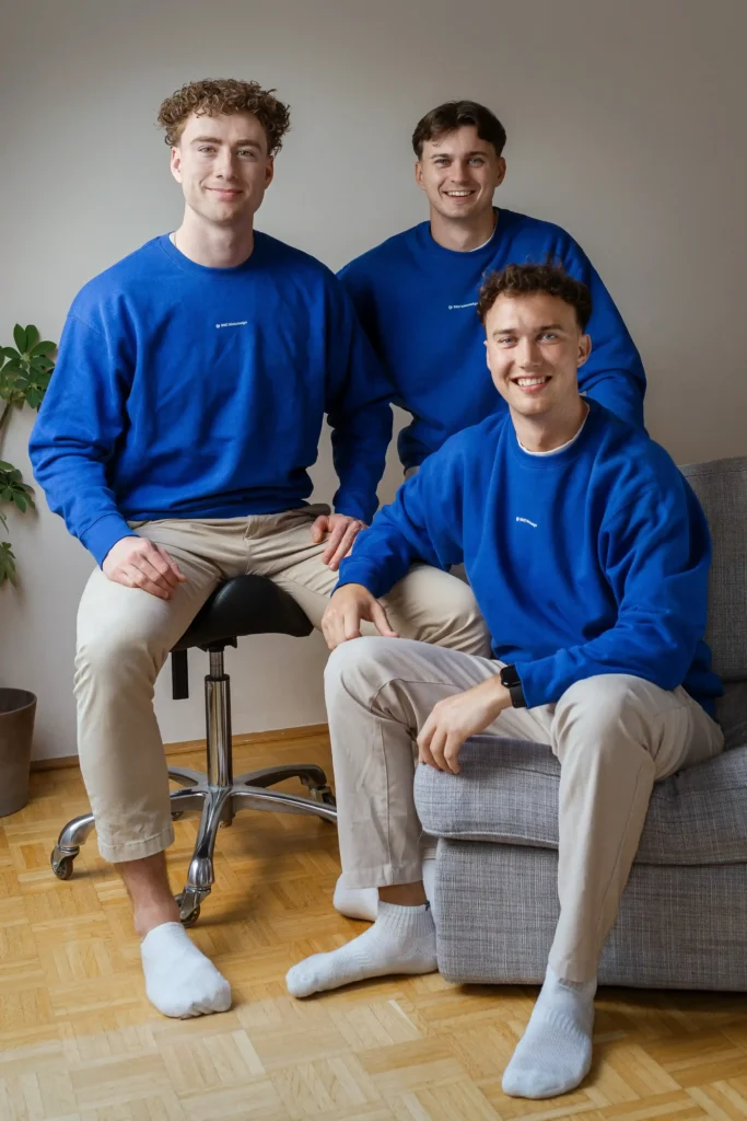 Gruppenfoto von drei jungen Männern in blauen Pullovern, zwei sitzend auf einem Sofa, einer stehend dahinter, vor hellem Hintergrund.