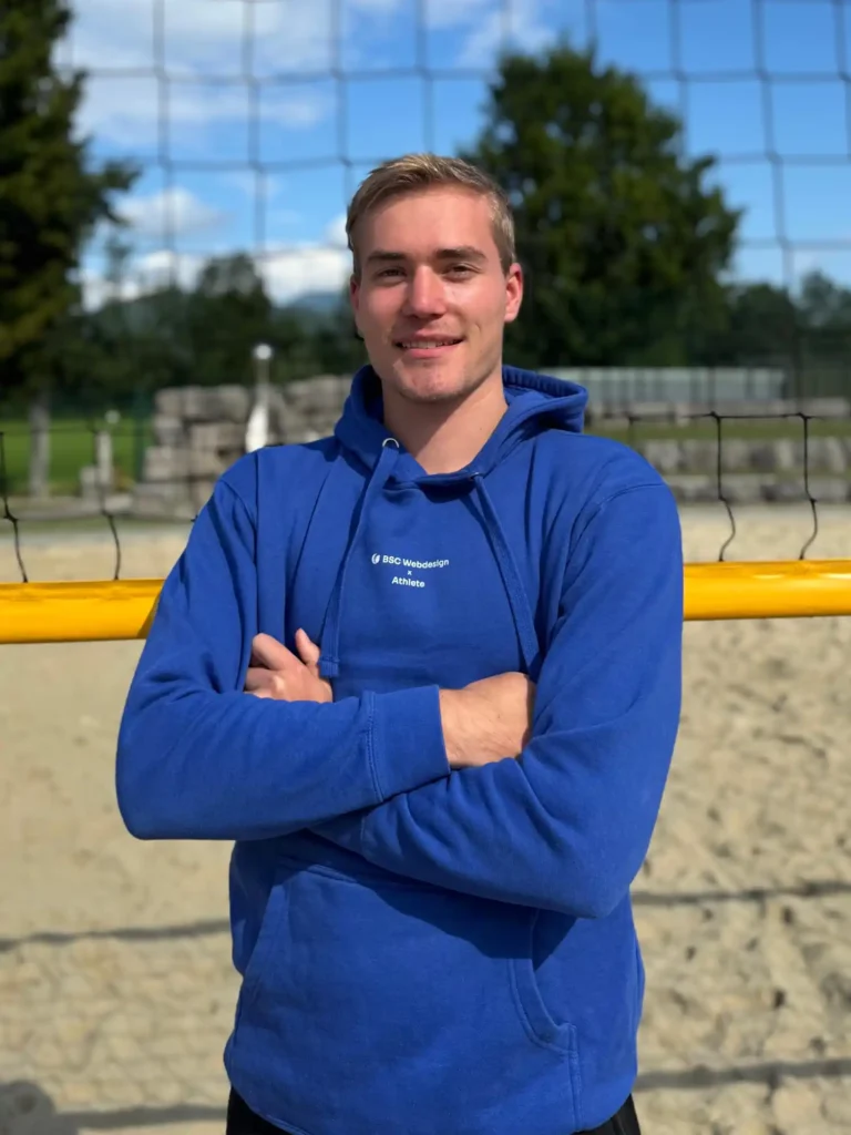 BSC Webdesign Athlete Daniel Ritzer mit blauem Hoodie am Beachvolleyballfeld bei Sonnenuntergang.
