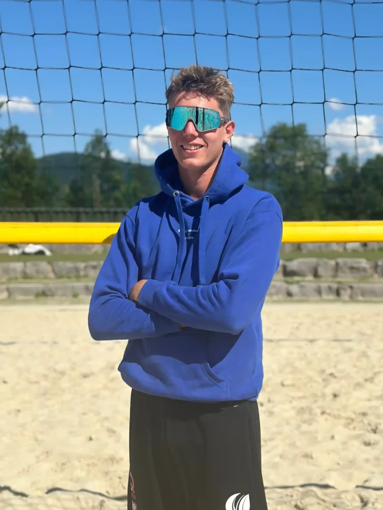 BSC Webdesign Athlete Tom Wallner mit blauem Hoodie am Beachvolleyballfeld bei Sonnenuntergang.