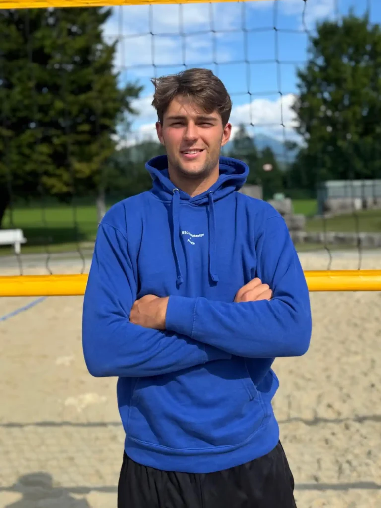 BSC Webdesign Athlete Xaver Stadlmann mit blauem Hoodie am Beachvolleyballfeld bei Sonnenuntergang.